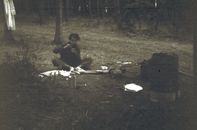Vintage black-and-white photo of a man seated on grass beside a makeshift campsite at dusk. Notebook and tin can beside him s...