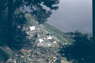 Aerial view of lakeside camping site with tents and scattered gear on grassy, forested shore. Reflective water and a small bo...