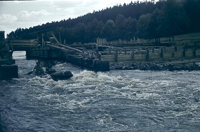 Industrial waterway with concrete-lined channel and wooden bridge structure, likely a hydroelectric dam or floodgate. Turbule...