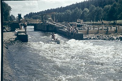 Historic wooden dam with water turbine and spillway in operation, mid-20th century. Two figures—one standing on platform, one...