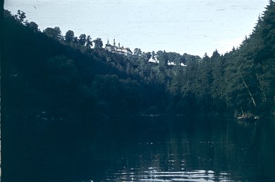 Vintage black-and-white photo of a lakeside scene featuring a grand, multi-domed building perched atop a forested hill. The r...
