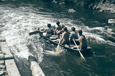 Six men in traditional wooden rowboats navigate turbulent waters, likely a river or rapid. Mid-20th century attire suggests o...