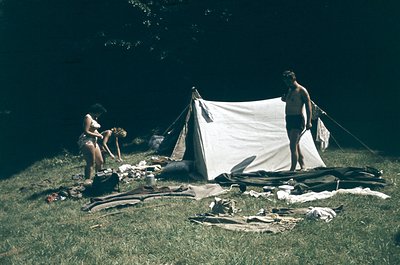 Vintage camping scene with two individuals setting up a white canvas tent in a grassy, wooded area. One person kneels organiz...