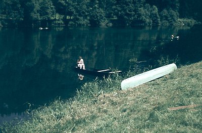 Vintage black-and-white photo of a lone individual seated on a docked canoe beside a calm lake, surrounded by dense forest. R...