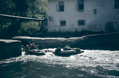 Vintage black-and-white photo of two individuals rafting on turbulent river rapids, likely in alpine terrain. Wooden rafts wi...