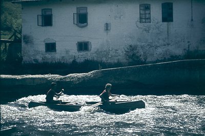 Two individuals in a narrow wooden boat navigate turbulent river waters, likely for fishing or transport. Behind them, a weat...