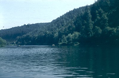 Vintage aerial view of a serene lake bordered by dense forested hills. Two small boats glide across calm waters, reflecting m...