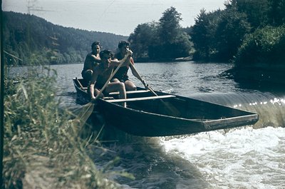 Vintage black-and-white boat scene with two adults and a child rowing a narrow wooden skiff on a calm river, surrounded by de...