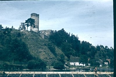 Vintage sepia-toned photo of a medieval stone tower perched atop a forested hillside, overlooking a serene lake. Remnants of ...