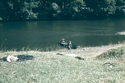 Vintage lakeside scene with two individuals seated on a wooden bench near a shallow water’s edge. Overgrown grass and a small...