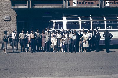 Group of 20+ people posing in front of a vintage bus (1960s-70s) near a brick building with signs "Vinárna" and "Resfri". Urb...