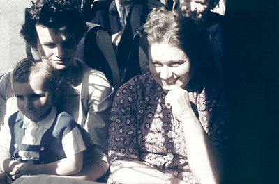 Vintage sepia-toned photo of a family group in mid-20th century attire, likely 1940s–1950s. A woman in patterned blouse and a...