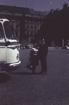 Mid-20th century urban scene featuring a vintage white taxi (likely a Type 2) parked on cobblestone streets. A man in a dark ...