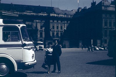 Vintage urban scene featuring a man and woman in mid-20th-century attire (1950s-60s) standing near a vintage bus in a Europea...