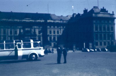 Vintage urban street scene featuring a mid-20th century tram or trolleybus in motion, passing a pedestrian in dark coat. Surr...