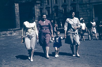 A family of four walks through a European city street in the 1950s, holding hands. The woman in the center wears a floral dre...