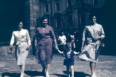 Three women and a child walk hand-in-hand on a European street, 1950s. Architectural details include ornate balconies and wro...