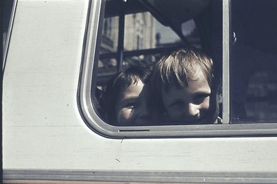 Two children peer out of a vintage bus window, framed by the metal sill. Their expressions suggest curiosity or contemplation...