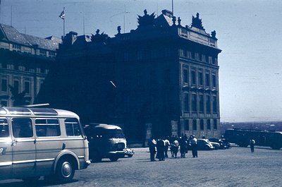 Vintage black-and-white street scene featuring a grand, ornate building with classical architecture—crenellated roofline, arc...