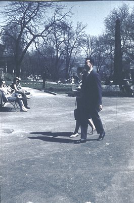Black-and-white photo of two men in formal attire (dark suits, ties) walking in a park. One man holds a cigarette. Leisurely ...