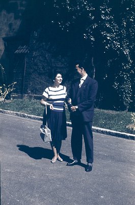 Couple in mid-20th century attire—woman in striped blouse & knee-length skirt, man in suit—posing outdoors on paved path. Lus...
