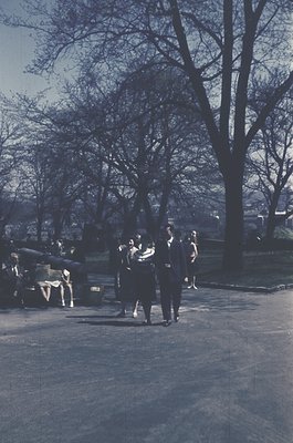 Vintage sepia-toned park scene with 1950s-60s fashion: group of men in suits and women in dresses walking near a bench. Leave...