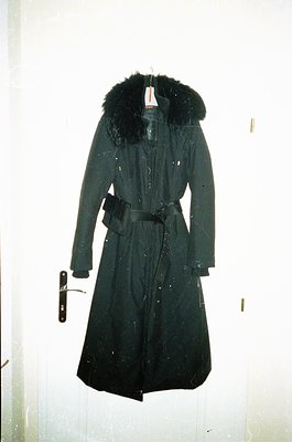 Mid-length black wool trench coat with fur-lined hood, belted waist, and visible price tag. Classic 1960s–1970s design, likel...