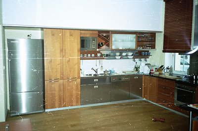 Mid-century kitchen with wood-paneled cabinets, stainless steel fridge, and retro appliances. Open shelving holds glassware a...