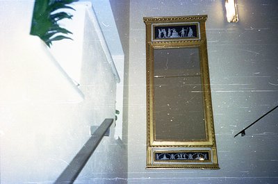 Vintage gold-framed mirror with decorative relief at base, mounted on light marble wall. Reflected image shows a staircase wi...