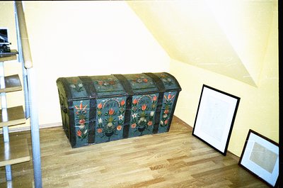 Vintage wooden trunk with floral motifs in deep blues, reds, and golds, placed on wooden flooring. Empty frames and metal sta...