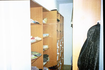 Vintage wooden closet with open shelves holding folded clothes, shoes, and plastic-wrapped items. Lighting suggests indoor re...