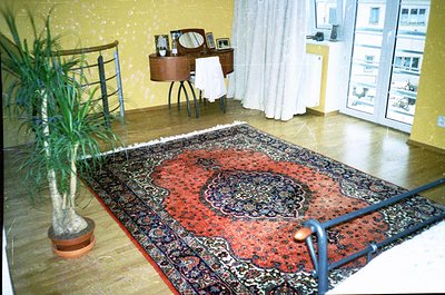 Mid-century modern bedroom with vintage furnishings: ornate red/black patterned rug, wooden vanity with mirror, and potted pl...