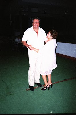 Couple dancing in a dimly lit indoor venue, likely a 1980s/1990s social event. Man in white suit, woman in sleeveless white d...