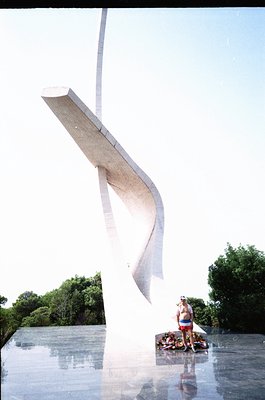 Modernist concrete sculpture resembling a wave or sail, anchored in reflective water. A person in swimwear poses beside it, s...
