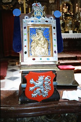 Religious ornate clock featuring a gold-toned icon of the Virgin Mary and Child, framed in silver with blue enamel accents. B...
