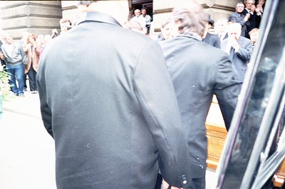 Blurred crowd scene featuring men in dark, formal attire (likely suits) entering a grand building with reflective glass doors...