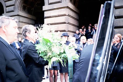 Formal gathering at an ornate stone building entrance, likely a funeral or memorial. Attendees in 1970s-era suits and dresses...