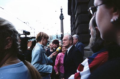 1970s-era public event near Nelson’s Column, London. Elderly man in suit speaking to crowd, surrounded by media (microphone, ...