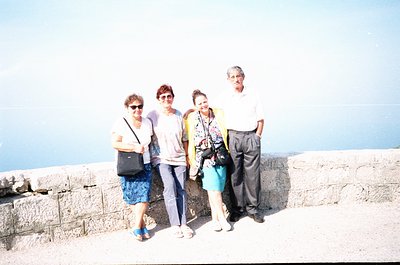 Four individuals pose on a stone wall overlooking a coastal horizon, likely 1990s–2000s. The woman on the left wears a crossb...