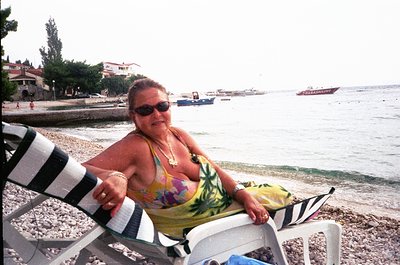 Vintage seaside portrait of a woman in a floral swimsuit, reclining on a striped beach chair by the shore. Small fishing boat...