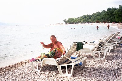 A relaxed individual sits on a pebble beach in a floral sarong, holding a woven bracelet, with lounge chairs and towels in a ...