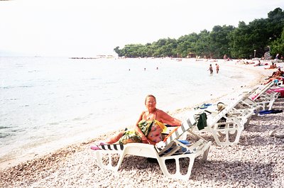 Two adults relax on vintage-style beach lounge chairs on a pebble shore, holding a striped towel. The turquoise sea and fores...