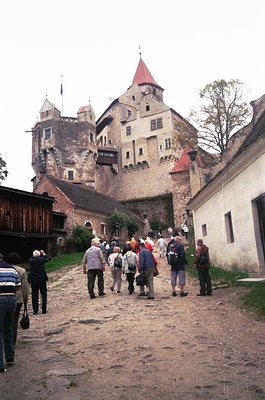 Medieval stone fortress with conical red-tiled towers dominates cobblestone courtyard. Tourists in casual 1990s attire explor...