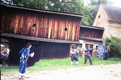 Historic reenactment scene featuring four individuals in 18th-century military attire—dark coats, white crossbelts, tricorn h...