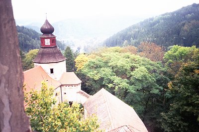 Historic castle tower with red-tiled conical roof and clock, surrounded by dense autumn foliage. Misty mountain backdrop sugg...