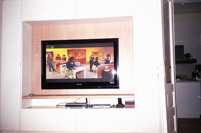 Flat-screen CRT TV mounted in recessed alcove, displaying a 1970s-style talk show with panelists and audience. Minimalist int...