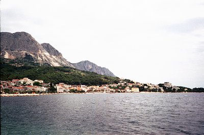Coastal town nestled between rugged mountains and deep blue sea, likely Mediterranean. Mid-20th century architecture with low...