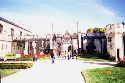 Historic wrought-iron gatehouse with ornate arches and glass conservatory wings flanking a sunlit courtyard. Red flowers and ...