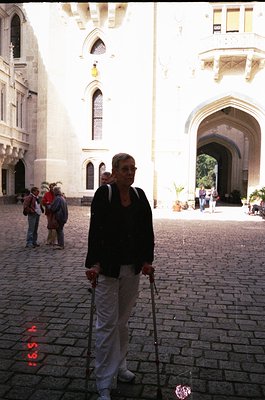 A woman in a dark sweater and light pants uses canes while walking in a historic courtyard. Gothic-style stone arches and poi...