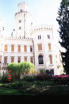Neoclassical-style castle with crenellated towers and arched windows, set against a clear sky. Lush garden with vibrant flowe...